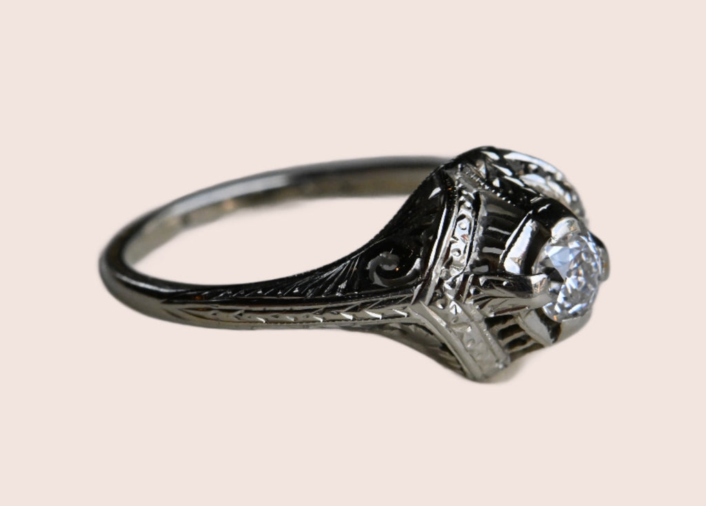 An Art Deco .25 carat Old European Cut diamond engagement ring with a gorgeous 18K white gold hand detailed mounting.  Ring sitting on neutral gray paper background.