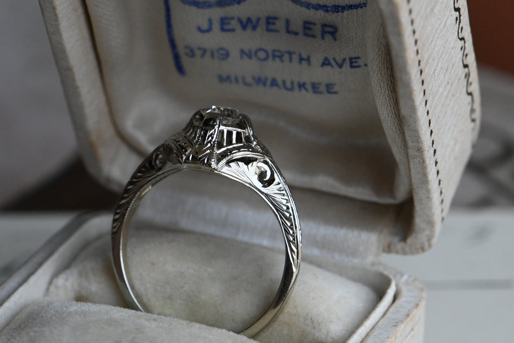 An Art Deco .25 carat Old European Cut diamond engagement ring with a gorgeous 18K white gold hand detailed mounting.  Ring sitting in cream colored antique ring box.
