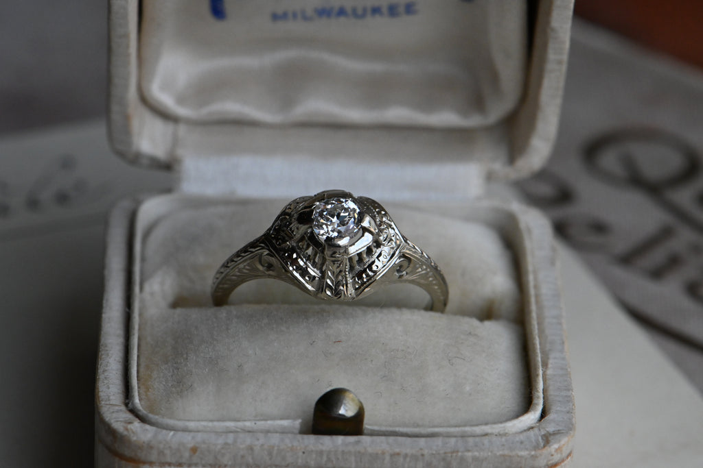 An Art Deco .25 carat Old European Cut diamond engagement ring with a gorgeous 18K white gold hand detailed mounting. Ring sitting in cream colored antique ring box.