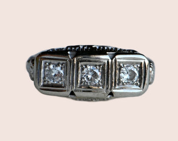 This Art Deco triple-diamond ring is fashioned in 14K white gold with the prettiest filigree and floral details. The shoulders have single-stem flowers - perhaps daisies! - leading to the head of the ring. Further filigree and floral flourishes on the side galleries make this a stunning ring from every angle! The old cut diamonds are bead set in square frames, flush to the setting and horizontally aligned. 