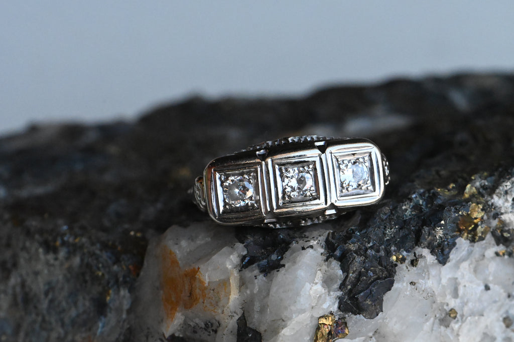 This Art Deco triple-diamond ring is fashioned in 14K white gold with the prettiest filigree and floral details. The shoulders have single-stem flowers - perhaps daisies! - leading to the head of the ring. Further filigree and floral flourishes on the side galleries make this a stunning ring from every angle! The old cut diamonds are bead set in square frames, flush to the setting and horizontally aligned. 