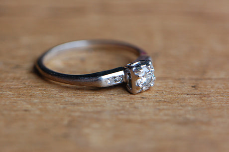 A classic late 1930s diamond solitaire engagement ring set in platinum with old cut diamonds. The diamond is set with five diamonds - a primary, Old European Cut center and flanking pairs of single cuts. The Old Euro is set in a square head with bead prongs and the single cuts are channel set. Ring photographed laying on a piece of grainy wood.