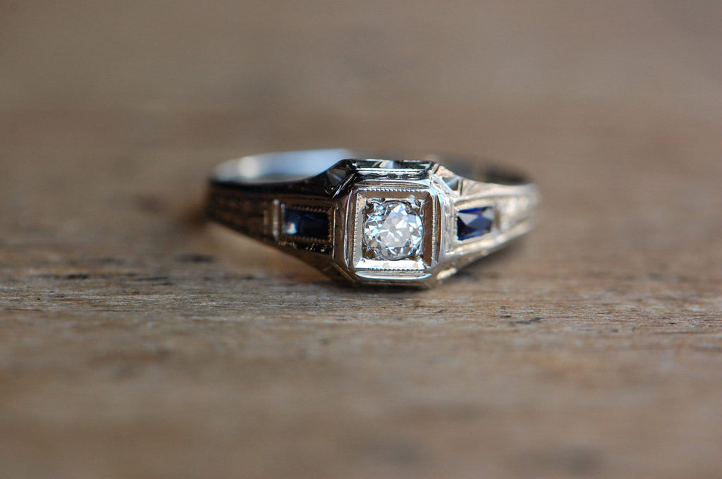 A handsome ca. 1920s Art Deco ring in 20K white gold (!) with an Old European Cut diamond primary and flanking sapphire baguettes. The hand-engraved details are exceptional and appear throughout the mounting. Split leaf motifs up the shoulders, milgrain around the baguettes, and etchings around the head are a few or our favorite accents. 