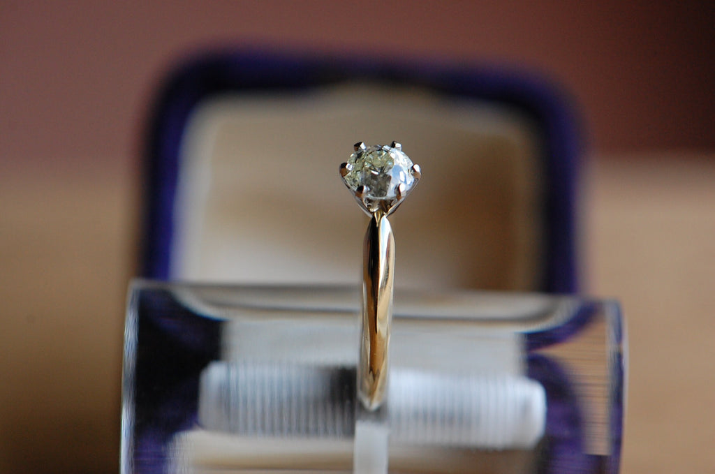 close up of solitaire diamond ring from the side angle. The ring is being held by a clear acrylic cylinder with blue vintage ring box out of focus in background.