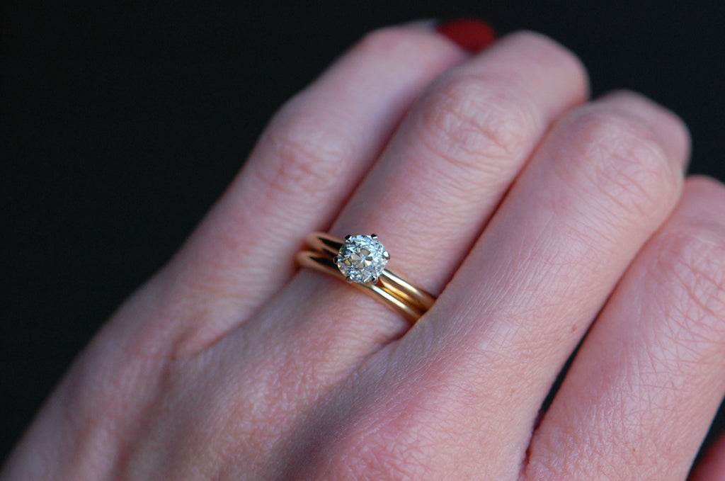 close up photo of solitaire diamond ring being modeled on ring finger. Diamond is set in a platinum 6 prong head with a 14k gold knife edge band. Ring is paired with another gold wedding band. Black background.