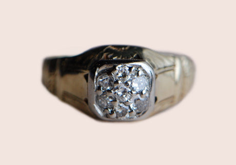 A Late Art Deco gentleman's diamond cluster ring with a magnificent hand-engraved mounting. Seven Old European Cut diamonds are pavé set in a tight cluster within a square head. The yellow gold shank is substantial (nearly 7 grams), comfortable on the hand, and beautifully engraved up the shoulders and around the side gallery. Ring close up of face of ring. Photographed against a mottled gray pink color.