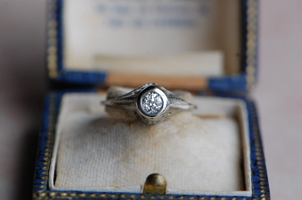 An Art Deco .38 carat Old European Cut diamond engagement ring with a 14K white gold mounting and hand-engraved details. The diamond is bezel set at the summit of the ring, with the decorative shoulders descending steeply from the round head. Ring in antique ring box.