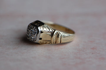 A Late Art Deco gentleman's diamond cluster ring with a magnificent hand-engraved mounting. Seven Old European Cut diamonds are pavé set in a tight cluster within a square head.  The yellow gold shank is substantial (nearly 7 grams), comfortable on the hand, and beautifully engraved up the shoulders and around the side gallery. Ring close up of side shoulder. Photographed against a mottled gray pink color.
