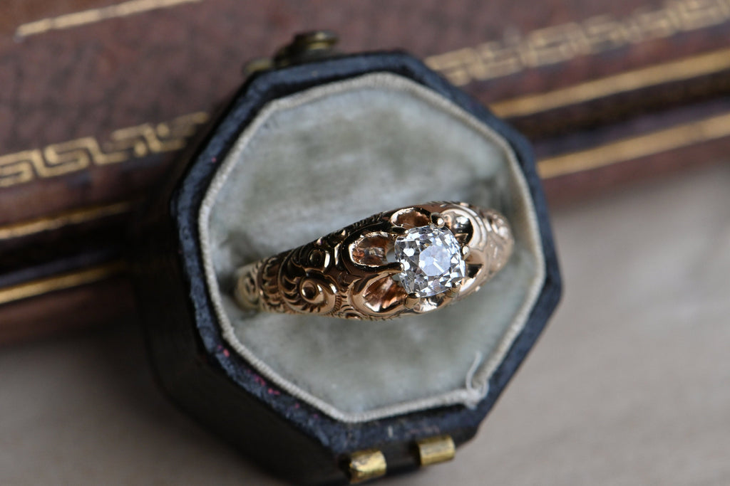 A 14K Victorian diamond engagement ring set with a plump Old Mine Cut diamond, just shy of 1/2 carat. The belcher set mounting features deep, dense chasing up the shoulders. The prongs are continuations of the shoulders themselves, reaching to grip the cushion-shaped diamond in a low profile. From above, the negative space between the prongs gives the head of the ring a petaled-look. Close up of head of ring with a squarish shaped diamond. Ring photographed inside of fabric lined antique ring box.