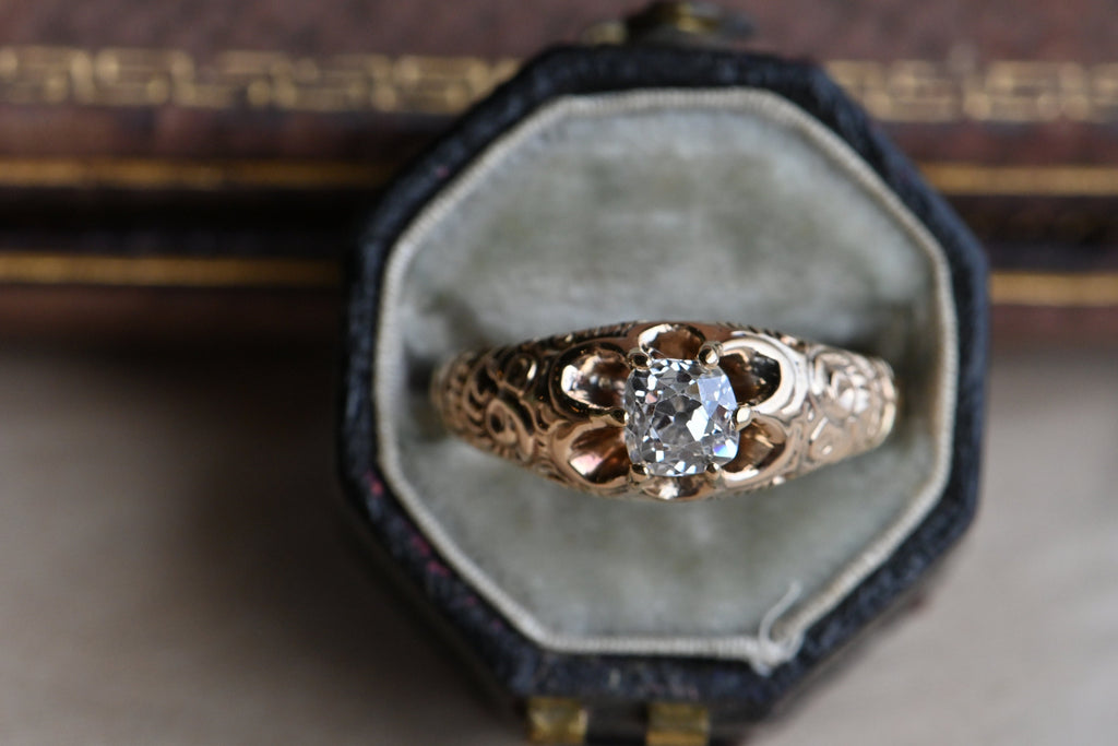 A 14K Victorian diamond engagement ring set with a plump Old Mine Cut diamond, just shy of 1/2 carat. The belcher set mounting features deep, dense chasing up the shoulders. The prongs are continuations of the shoulders themselves, reaching to grip the cushion-shaped diamond in a low profile. From above, the negative space between the prongs gives the head of the ring a petaled-look. Close up of head of ring with a squarish shaped diamond. Ring photographed inside of fabric lined antique ring box.