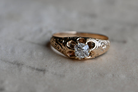 A 14K Victorian diamond engagement ring set with a plump Old Mine Cut diamond, just shy of 1/2 carat. The belcher set mounting features deep, dense chasing up the shoulders. The prongs are continuations of the shoulders themselves, reaching to grip the cushion-shaped diamond in a low profile. From above, the negative space between the prongs gives the head of the ring a petaled-look.  Close up of head of ring with a squarish shaped diamond. Ring laying on top of textured gray colored paper.