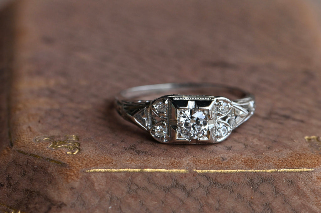 An Art Deco engagement ring with a .27 carat Old European Cut diamond tucked in an ornate mounting. There are so many things to appreciate here, not least of all the .27 carat Old European Cut center stone. This stone is tucked in a square head with beveled and notched details.