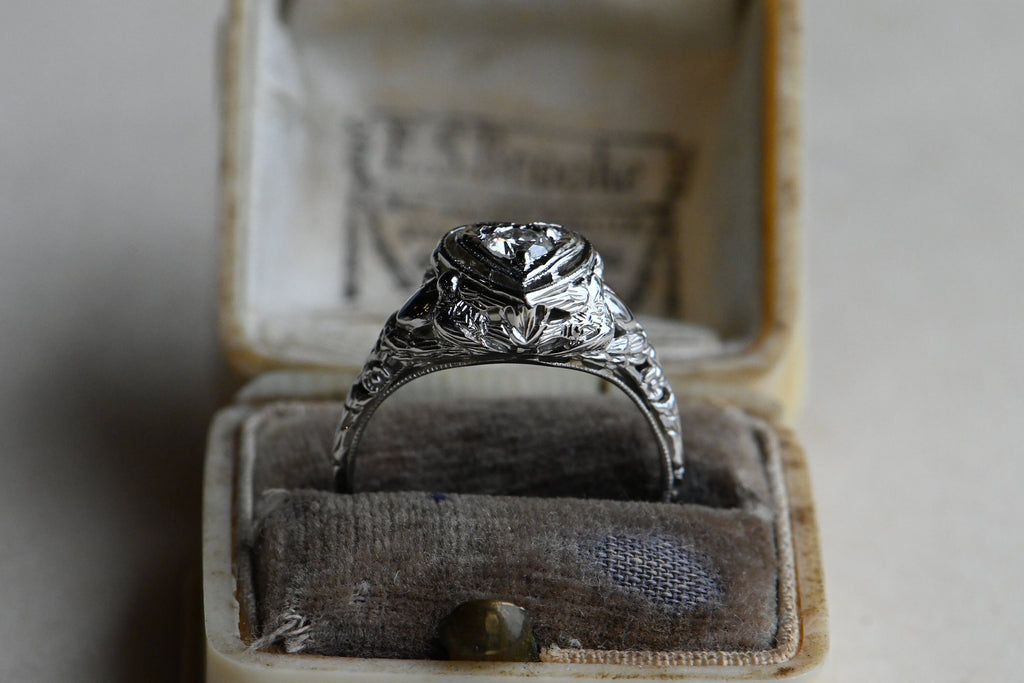 A romantic Art Deco filigree engagement ring with an Old European diamond illusion set in a heart-shaped head. The diamond is set in a floating, heart-shaped head with milgrain detail on every edge. This ring looks like it houses a MUCH bigger diamond than it does. Ring inside gray and cream colored antique ring box.