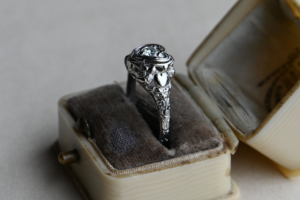 A romantic Art Deco filigree engagement ring with an Old European diamond illusion set in a heart-shaped head. The diamond is set in a floating, heart-shaped head with milgrain detail on every edge. This ring looks like it houses a MUCH bigger diamond than it does. Ring inside gray and cream colored antique ring box.