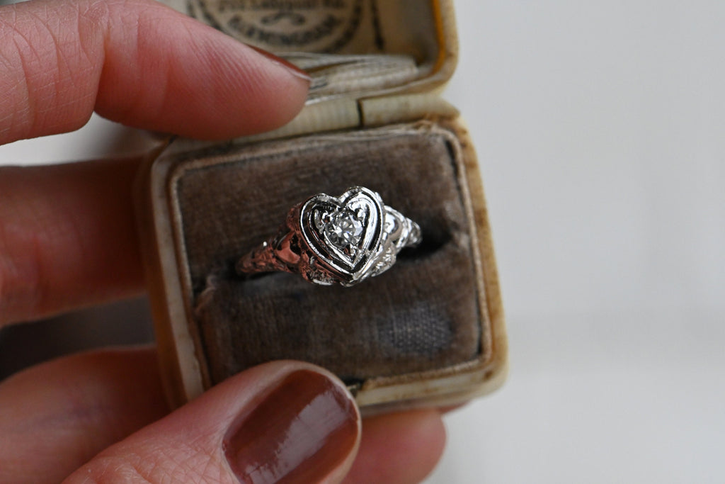 A romantic Art Deco filigree engagement ring with an Old European diamond illusion set in a heart-shaped head.  The diamond is set in a floating, heart-shaped head with milgrain detail on every edge. This ring looks like it houses a MUCH bigger diamond than it does. Ring inside antique ring box being held by female hand with rust colored nail polish.