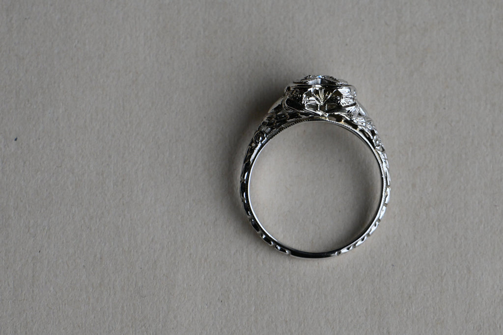 A romantic Art Deco filigree engagement ring with an Old European diamond illusion set in a heart-shaped head. The diamond is set in a floating, heart-shaped head with milgrain detail on every edge. This ring looks like it houses a MUCH bigger diamond than it does. Ring against neutral gray paper background.
