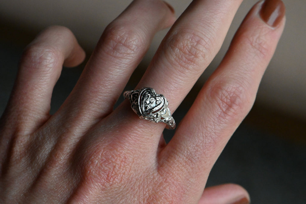 A romantic Art Deco filigree engagement ring with an Old European diamond illusion set in a heart-shaped head. The diamond is set in a floating, heart-shaped head with milgrain detail on every edge. This ring looks like it houses a MUCH bigger diamond than it does. Ring modeled on female hand with rust colored nail polish.