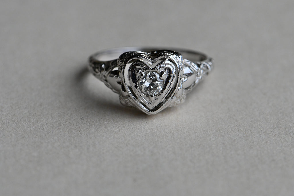 A romantic Art Deco filigree engagement ring with an Old European diamond illusion set in a heart-shaped head. The diamond is set in a floating, heart-shaped head with milgrain detail on every edge. This ring looks like it houses a MUCH bigger diamond than it does. Ring against neutral gray paper background.