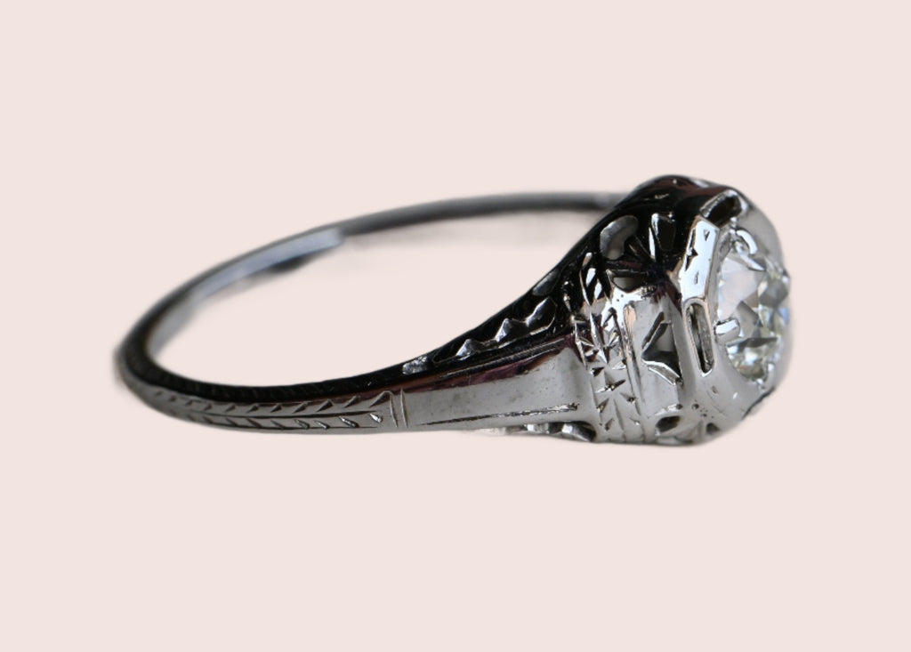 Art Deco engagement ring! This ring has hand-sawed filigree details and a juicy old mine cut feature stone. The 18K white gold mounting has engraved details on every millimeter, including bridal wreaths down the sides of the slim shank. Ring on gray paper background.