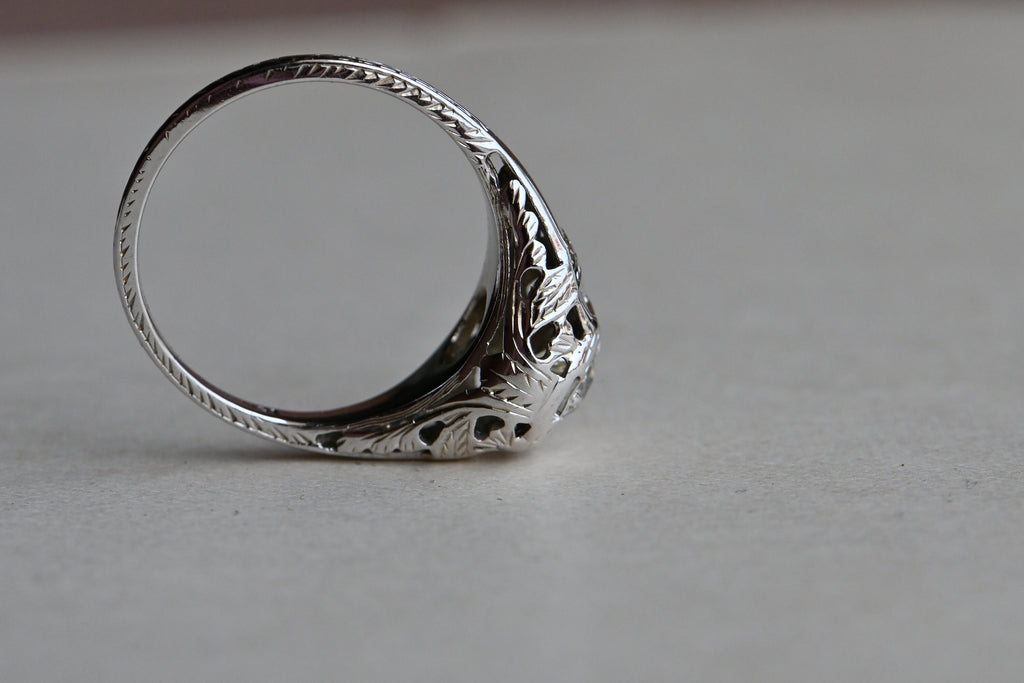 Art Deco engagement ring! This ring has hand-sawed filigree details and a juicy old mine cut feature stone. The 18K white gold mounting has engraved details on every millimeter, including bridal wreaths down the sides of the slim shank. Ring on gray paper background. Side profile in focus.