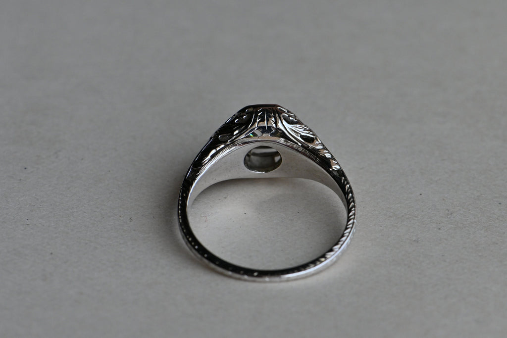 Art Deco engagement ring! This ring has hand-sawed filigree details and a juicy old mine cut feature stone. The 18K white gold mounting has engraved details on every millimeter, including bridal wreaths down the sides of the slim shank. Ring on gray paper background. Inside of ring in focus.