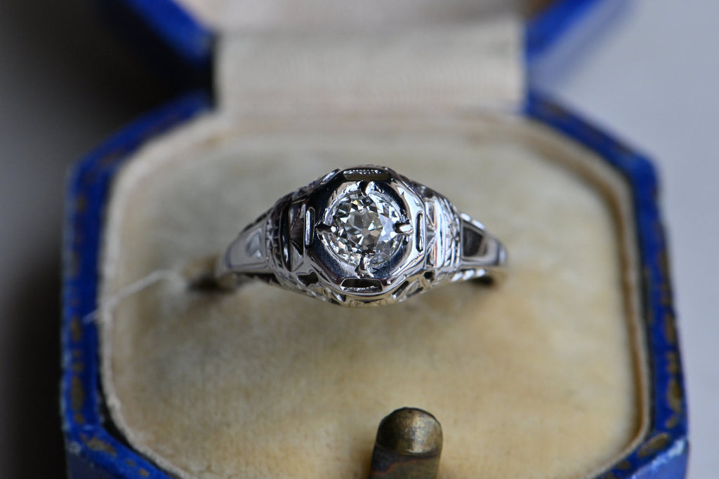 Art Deco engagement ring! This ring has hand-sawed filigree details and a juicy old mine cut feature stone. The 18K white gold mounting has engraved details on every millimeter, including bridal wreaths down the sides of the slim shank. Ring in antique ring box with cream and blue.
