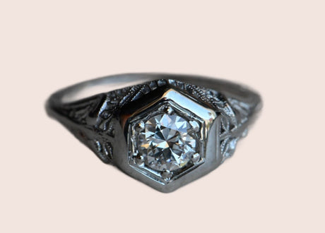 A classic Art Deco solitaire ring with a .56 carat Old European Cut diamond tucked in a romantic filigree mounting. The VS1/H diamond rests in an 18K white gold setting. Lacy filigree details up the shoulders. Ring sitting on gray paper.