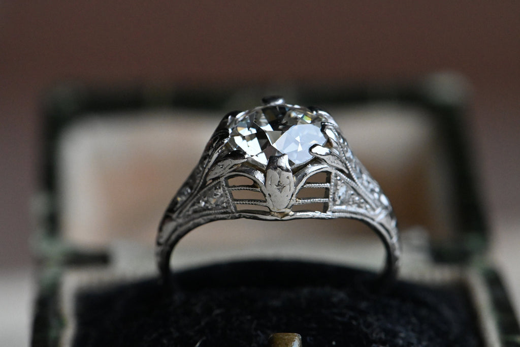 This ring has a platinum filigree mounting as fine as French lingerie (oh yes) with side gallery openwork and milgrained shoulders that are set with ten Old European Cut diamonds flanking the ès jolie Old European Cut Center diamond. Ring in black velvet box.