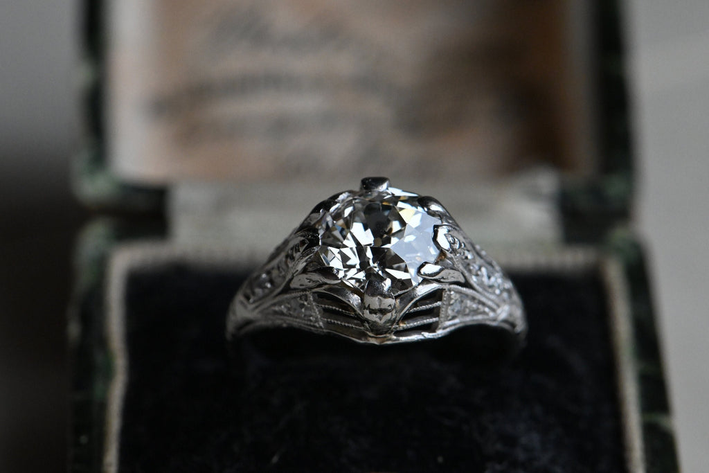  This ring has a platinum filigree mounting as fine as French lingerie (oh yes) with side gallery openwork and milgrained shoulders that are set with ten Old European Cut diamonds flanking the ès jolie Old European Cut Center diamond. Ring in black velvet box.