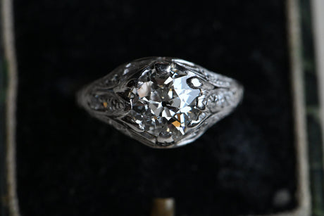This ring has a platinum filigree mounting as fine as French lingerie (oh yes) with side gallery openwork and milgrained shoulders that are set with ten Old European Cut diamonds flanking the ès jolie Old European Cut Center diamond. Ring on black velvet background.