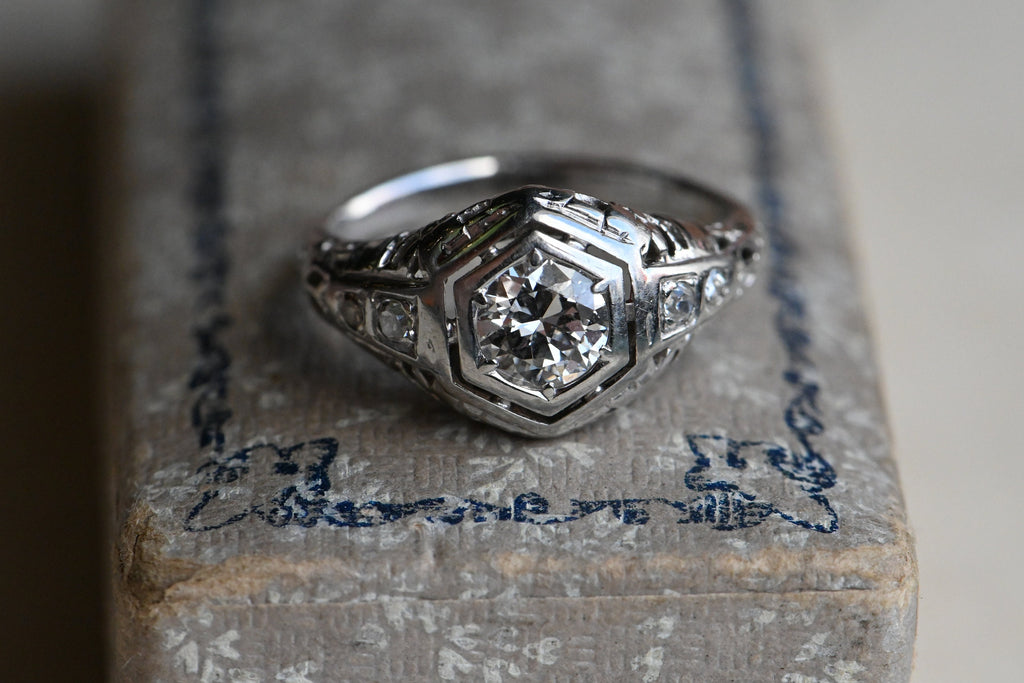 A classic Art Deco solitaire ring with a .45 carat Old European Cut diamond set in 18K white gold filigree.The diamond is flush-set in a floating, hexagonal head. This diamond is lively and crisp, graded VS2/G. Close up of ring  set on marbled  paper box  with blue.