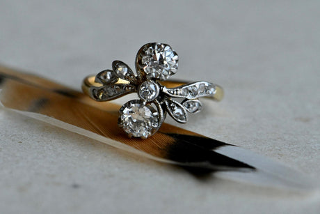 An elegant antique diamond toi et moi ring modeled in 18K yellow gold and platinum.  A pair of Old European Cut diamonds - a 1/3 carat each - are held vertically in the curves of a supple "S" frame, appearing almost to be tied on the finger like a sparkling bow. A third, smaller Old Euro rests between them at the center of the design. A sensual ring that flatters the finger. Close up of ring sitting on brown and black feather which is atop of gray colored paper.
