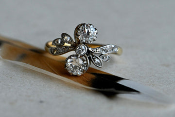 An elegant antique diamond toi et moi ring modeled in 18K yellow gold and platinum.  A pair of Old European Cut diamonds - a 1/3 carat each - are held vertically in the curves of a supple "S" frame, appearing almost to be tied on the finger like a sparkling bow. A third, smaller Old Euro rests between them at the center of the design. A sensual ring that flatters the finger. Close up of ring sitting on brown and black feather which is atop of gray colored paper.