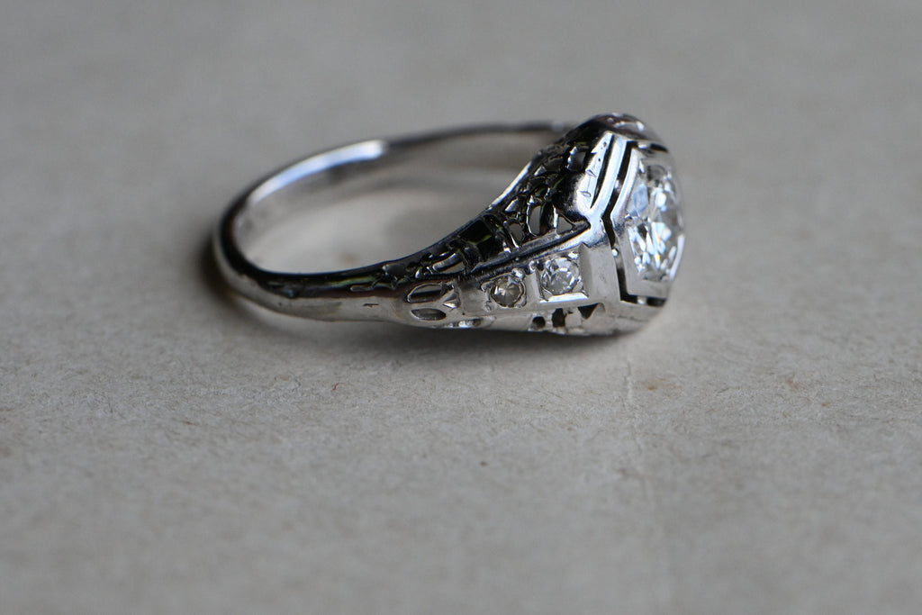 A classic Art Deco solitaire ring with a .45 carat Old European Cut diamond set in 18K white gold filigree.The diamond is flush-set in a floating, hexagonal head. This diamond is lively and crisp, graded VS2/G. Close up of shoulder of ring against neutral gray background.