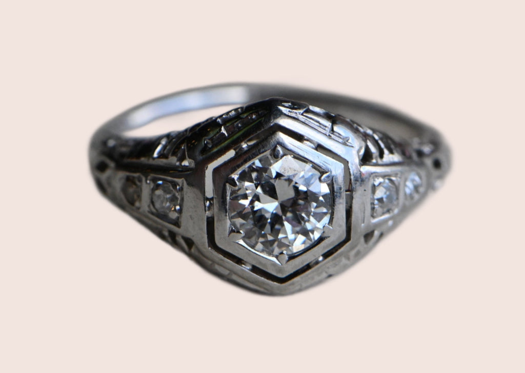 A classic Art Deco solitaire ring with a .45 carat Old European Cut diamond set in 18K white gold filigree.The diamond is flush-set in a floating, hexagonal head. This diamond is lively and crisp, graded VS2/G. Close up of face of ring against neutral gray background.