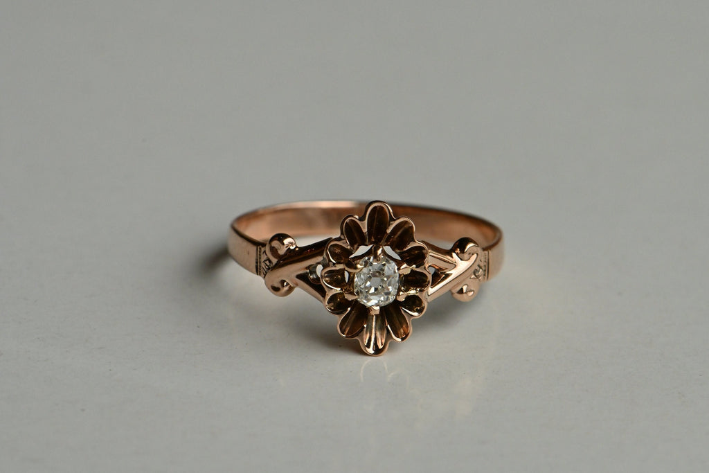  A modest yet plump 17 point old mine cut diamond is prong set at the center of a flared and elongated buttercup mounting. The head reminds me of a little tartlet pan! The shoulders are split with a sweet crossover pattern at the shank. Blushing 14K rose gold.  Ring photographed dating on top of gray colored paper.