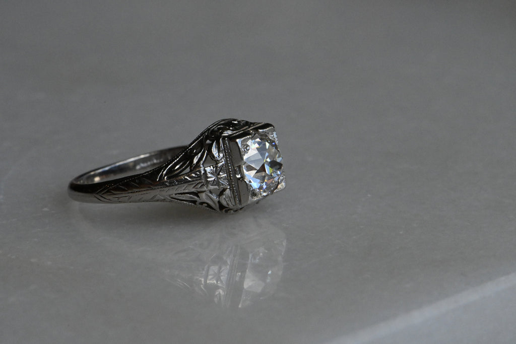 1920s Art Deco .85 carat diamond solitaire ring neatly modeled in platinum. The ring features a juicy Old European Cut diamond set in a square head. (VS1/H). The steep crown, small table, and petal-like facets of this diamond are beautiful markers of the antique cutting style. The remarkable setting is hand-carved and engraved with tendrils, leaves, and flowers. Ring close up against neutral background.  