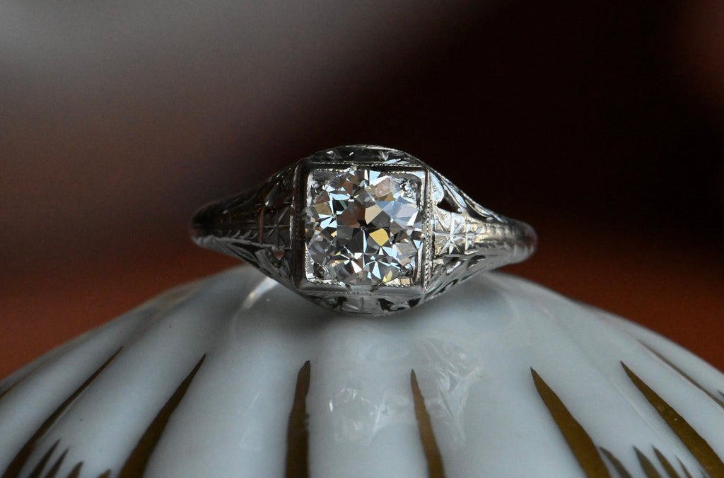 1920s Art Deco .85 carat diamond solitaire ring neatly modeled in platinum. The ring features a juicy Old European Cut diamond set in a square head. (VS1/H). The steep crown, small table, and petal-like facets of this diamond are beautiful markers of the antique cutting style. The remarkable setting is hand-carved and engraved with tendrils, leaves, and flowers. Ring  frontal close up on porcelain shell.