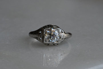 1920s Art Deco .85 carat diamond solitaire ring neatly modeled in platinum. The ring features a juicy Old European Cut diamond set in a square head. (VS1/H). The steep crown, small table, and petal-like facets of this diamond are beautiful markers of the antique cutting style. The remarkable setting is hand-carved and engraved with tendrils, leaves, and flowers. Ring close up against neutral background.