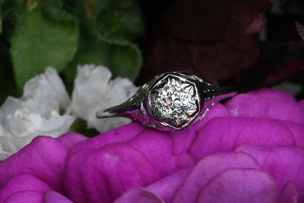 An Art Deco 18K diamond engagement ring with an Old European Cut diamond tucked in a hexagonal head.  Close up of head of ring on fascia flower petals with white flowers in background.