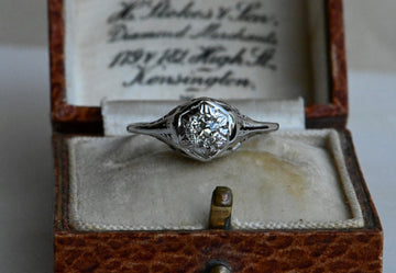 An Art Deco 18K diamond engagement ring with an Old European Cut diamond tucked in a hexagonal head. Ring sitting on old English vintage ring box.