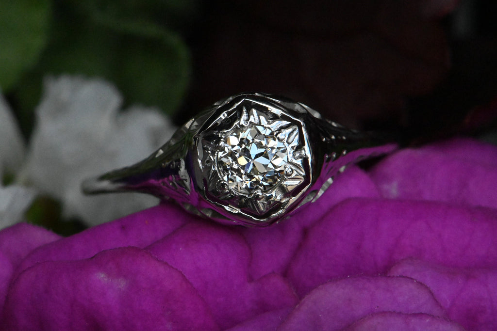 An Art Deco 18K diamond engagement ring with an Old European Cut diamond tucked in a hexagonal head. Close up of ring sitting askew on top of fuscia flower petals.