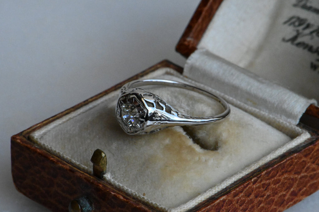 An Art Deco 18K diamond engagement ring with an Old European Cut diamond tucked in a hexagonal head.  Ring laying inside an antique ring box with shoulder of ring in focus.