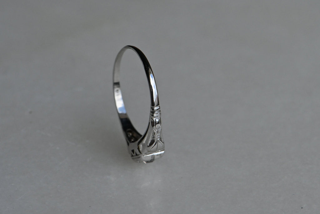 A dainty and understated 1930s ring with a .20 carat Old European Diamond in 14K white gold. The round diamond has a darling, tiny table and long, kite-shaped crown facets. If offers a bright, consistent sparkle from its perch in a notched, square-shaped head. The shoulders have pretty cast details reminiscent of bows or gathered fabric. Milgrain detail throughout. Ring photographed positioned on its side on top of white colored reflective surface.