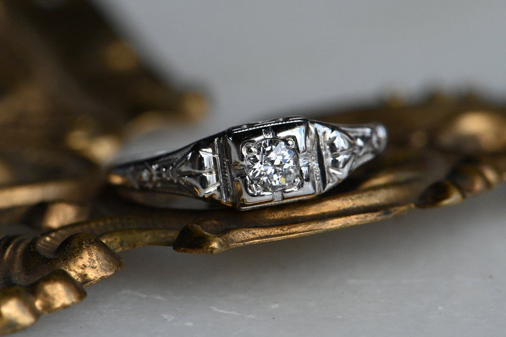 A dainty and understated 1930s ring with a .20 carat Old European Diamond in 14K white gold. The round diamond has a darling, tiny table and long, kite-shaped crown facets. If offers a bright, consistent sparkle from its perch in a notched, square-shaped head. The shoulders have pretty cast details reminiscent of bows or gathered fabric. Milgrain detail throughout.  Front of ring photographed on top of brass colored metalwork. White colored background.