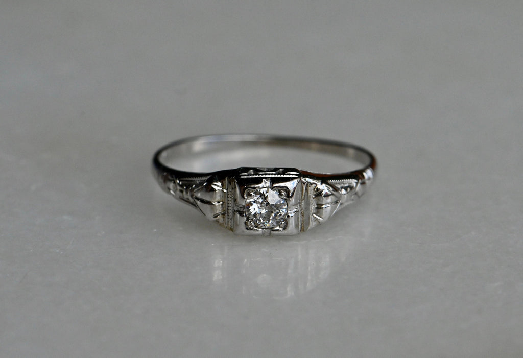 A dainty and understated 1930s ring with a .20 carat Old European Diamond in 14K white gold. The round diamond has a darling, tiny table and long, kite-shaped crown facets. If offers a bright, consistent sparkle from its perch in a notched, square-shaped head. The shoulders have pretty cast details reminiscent of bows or gathered fabric. Milgrain detail throughout. Ring photographed laying on top of white colored reflective surface.